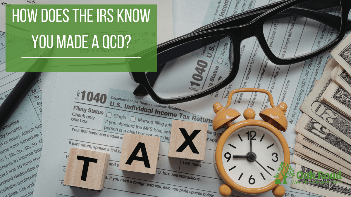 Overhead photograph of a tax planning desk set with a 2021 Form 1040, glasses, wooden blocks spelling "TAX", a vintage orange alarm clock, and a stack of money. A green overlay box asks: "HOW DOES THE IRS KNOW YOU MADE A QCD?". Includes the Oak Road Wealth Management logo.