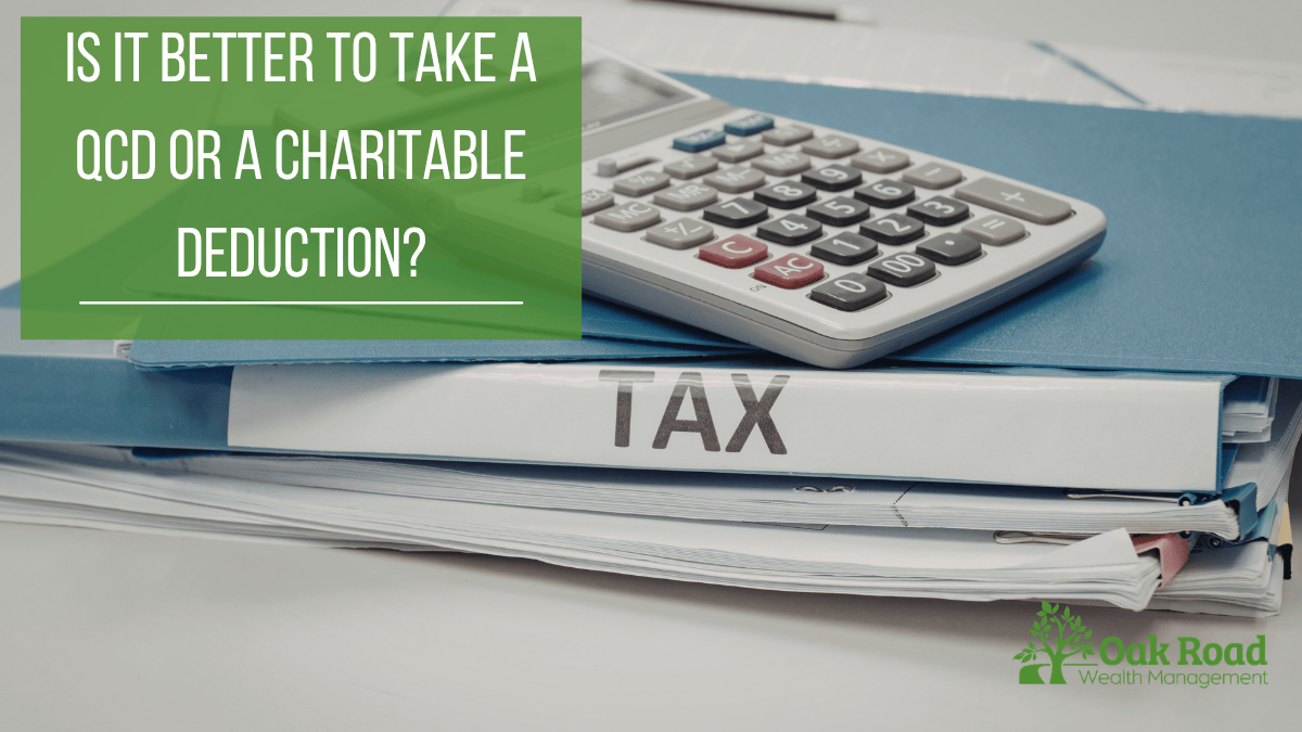 Detailed photograph of a stack of office folders and a calculator on a desk. The spine of a blue folder clearly displays the word 'TAX'. Overlaid in a green banner is the text: 'Is it better to take a Qualified Charitable Distribution (QCD) or a charitable deduction?' The Oak Road Wealth Management tree logo is in the bottom corner.